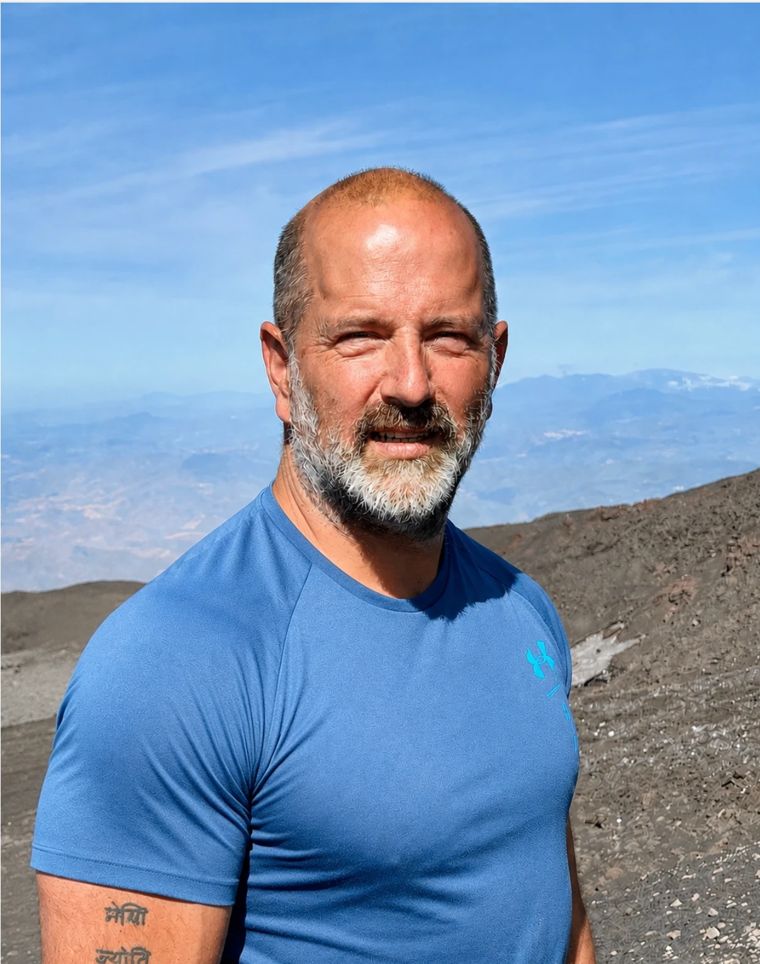 Will van Zyl outdoors on a mountain slope of dark volcanic ground, blue sky and distant hills behind him.